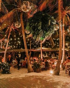 people are sitting at tables under palm trees and lights on the beach in front of them