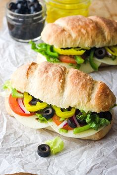 two sub sandwiches sitting on top of paper next to some olives and lettuce