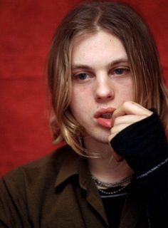 a young man with long hair wearing a black shirt and holding his hand to his mouth