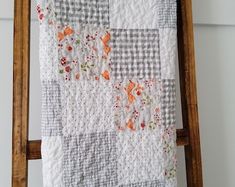 a quilt hanging on a ladder in front of a white wall with an orange and gray flowered design