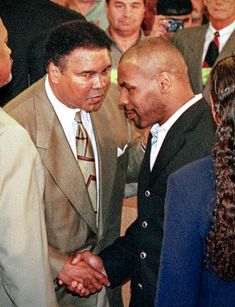 two men shaking hands in front of a group of people and one is wearing a suit