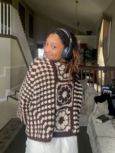 a woman with headphones on standing in front of a couch wearing a brown and white sweater