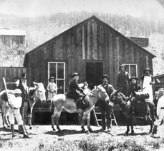 an old black and white photo of people with horses in front of a cabin on the side of a mountain