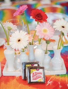 several white vases with flowers in them on a colorful table cloth and place setting