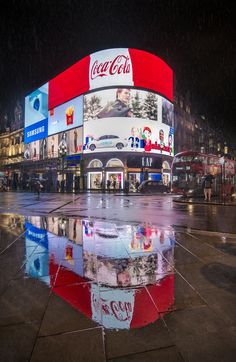 the reflection of a coca - cola building in a puddle on a city street at night