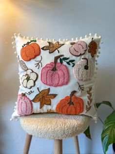 a white pillow with pumpkins and leaves on it sitting on a stool next to a potted plant