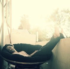 a man laying on top of a wooden chair next to a glass wall with sunlight coming through the window