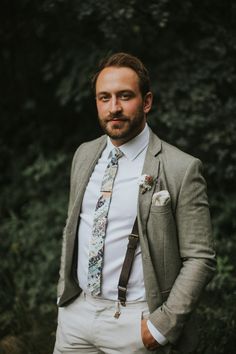 a man wearing a suit and tie standing in front of some green trees with his hands in his pockets