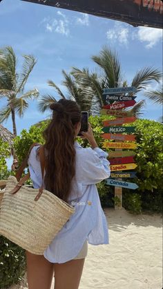 a woman is standing on the beach taking a photo with her cell phone and holding a straw bag