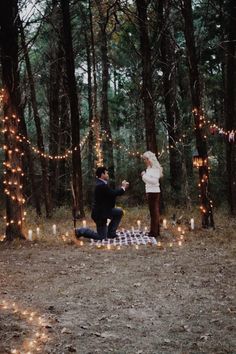 a man kneeling down next to a woman on a blanket in the woods surrounded by lights