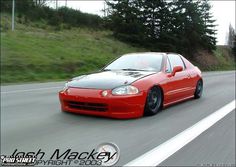 a red car driving down the road with trees in the backgrouund and grass behind it