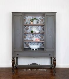 an old china cabinet painted in gray with flowers on the top and shelves above it