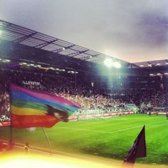 a rainbow flag is in the middle of a soccer field as people watch from the stands