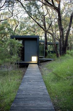 a wooden walkway leading to a small black structure in the middle of some grass and trees