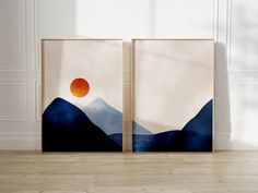 two framed pictures with mountains and a red sun in the middle, on a wooden floor