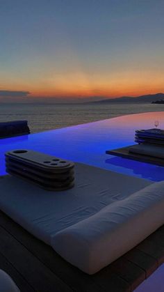 an outdoor swimming pool with lounge chairs overlooking the ocean at sunset or sunrise, in front of a blue and orange sky