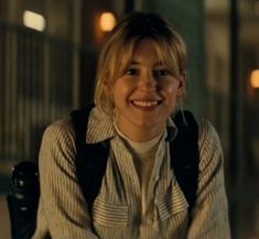 a woman with blonde hair smiling and wearing a backpack in front of a building at night