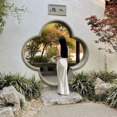 a woman standing in front of a mirror with plants and rocks around her, looking into it