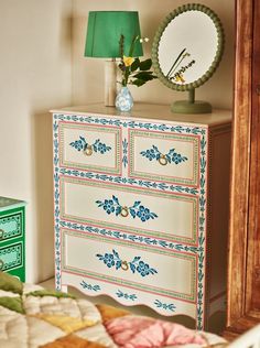 a white dresser with blue flowers on it and a green lamp next to the dresser