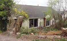 a house that is next to some trees in the grass and bushes on the side of the road