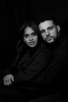 a man and woman sitting next to each other on a black background with their arms around each other