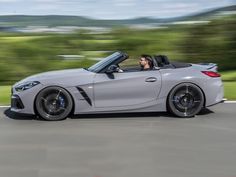 a man driving a silver convertible car down a curvy road with trees in the background