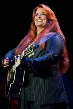 a woman with long red hair holding a guitar and smiling at the camera while standing in front of a microphone