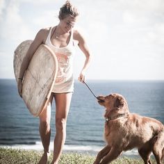 a woman walking her dog on a leash with a surfboard in its mouth and the ocean in the background