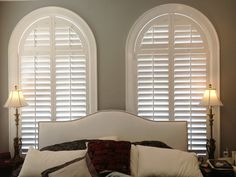 two arched windows with white shutters above a bed in front of pillows and lamps