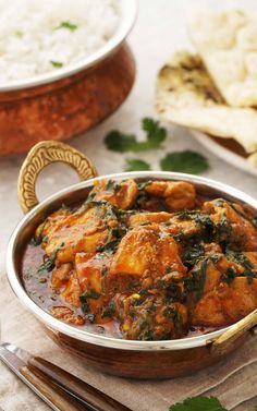 a metal bowl filled with chicken and spinach curry next to rice on a table