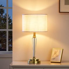 a lamp on a table next to a window with a picture frame and framed pictures