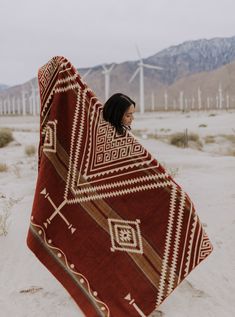 a woman is wrapped up in a blanket on the beach with wind mills behind her