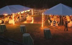 a group of people sitting at tables under white tents with string lights on the ground