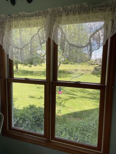 two windows with white curtains in the middle of a green yard and trees outside them