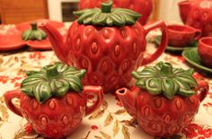 red ceramic strawberry teapots with matching cups and saucers on a floral tablecloth