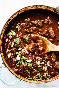 a pot filled with meat and vegetables on top of a white tablecloth next to a wooden spoon