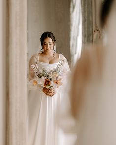 a woman standing in front of a mirror wearing a white dress and holding a bouquet