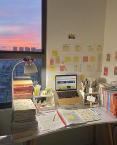 a desk with a laptop, books and papers on it in front of a window