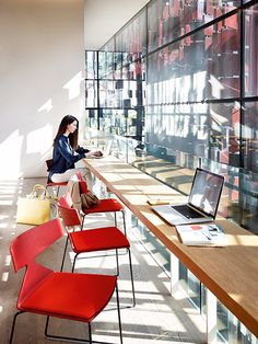 a woman sitting on a chair in front of a desk with a laptop and red chairs