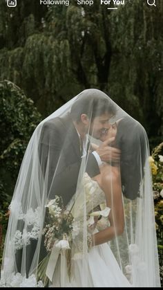 a bride and groom kissing under a veil in front of some trees on their wedding day