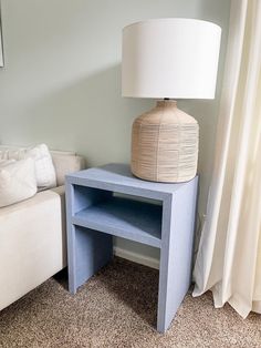 a blue table with a white lamp on it in front of a couch and window