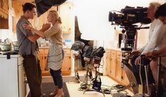 two men and a woman standing in a kitchen with camera equipment on the counter next to them