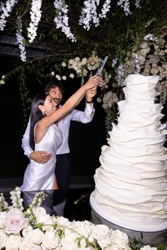 a man and woman standing next to a white wedding cake with flowers on the side