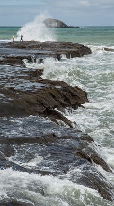 two people standing on rocks near the ocean with waves crashing against them and one person in yellow jacket