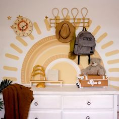 a white dresser topped with a teddy bear next to a wall mounted clock and hat rack