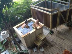 two people in a wooden hot tub on a deck next to some trees and plants