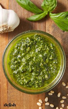 a glass bowl filled with pesto sauce next to garlic and green leaves on a wooden table