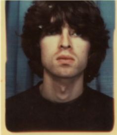 a man with long hair and black shirt in front of a blue curtain looking at the camera