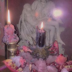 two candles sitting on top of a table next to some rocks and flowers in front of a statue