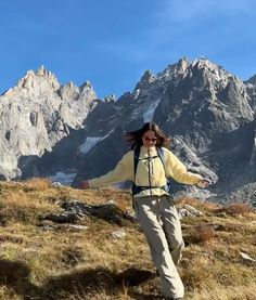 a woman standing on top of a grass covered field next to tall mountain peaks with her arms outstretched